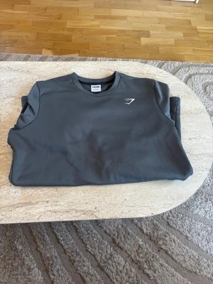 Mörkgrå sweatshirt från Gymshark - Snygg mörkgrå sweatshirt från Gymshark med rund halsringning och diskret vitt logotryck på bröstet. Tillverkad i ett mjukt och funktionellt material som passar perfekt till träning eller chill. Enkel och stilren design som är lätt att matcha.