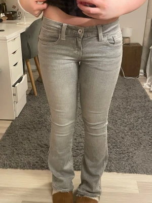 Grå lågmidjade bootcut jeans  - Säljer ett par ljusgrå bootcut jeans med snygga broderade detaljer på bakfickorna. Jeansen har klassisk femficksmodell, normal midja och är tillverkade i stretchigt denimtyg för skön passform. Perfekta till sneakers eller boots.