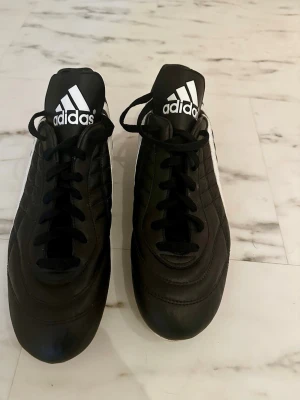 Adidas klassiska svarta fotbollsskor - Säljer ett par svarta Adidas fotbollsskor med vita ränder på sidorna och klassisk design. Skorna har fasta dobbar och är tillverkade i syntetmaterial med snörning framtill. Perfekta för matcher på gräsplan. Adidas-logga på plösen