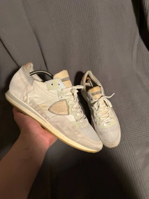  Philippe Model - Snygga beige sneakers från Philippe Model med detaljer i mocka och textil. Skorna har vita snören, ljusgula sulor och en ikonisk sköldlogga på sidan. Färgskalan är neutral med inslag av ljusgrått och krämvitt, perfekt för en clean och stilren look.