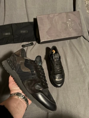 Valentino  - Snygga sneakers från Valentino Garavani i svart och mörkgrå camo. Skorna har en mix av mocka, läder och textil, samt coola nitar på hälen. Tjock sula och klassisk låg modell med svarta skosnören och logga på plösen.
