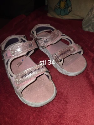 Rosa sandaler från Leaf stl 34 - Säljer ett par ljusrosa sandaler från Leaf i storlek 34. De har glittriga remmar med kardborrestängning och vit sula. Insidan är mjukt vadderad för extra komfort och sandalerna har en sportig look. Perfekta för sommaren!