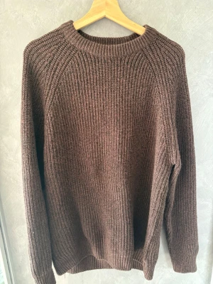 Brun stickad tröja i ullmix - Mysig brun stickad tröja med rund hals och långa ärmar. Tröjan har en grovstickad struktur och ribbade muddar vid ärmslut och nederkant. Perfekt för kyliga dagar och enkel att matcha med jeans eller kjol.