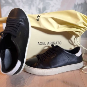 Axel Arigato sneakers i svart läder - Snygga svarta sneakers från Axel Arigato i läder med vit sula och vit detalj på hälen. Klassisk låg modell med guldfärgad logga på sidan och svarta skosnören. Perfekta för en clean och stilren look.