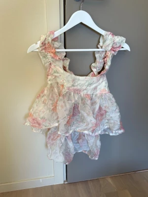 Blommig volangtopp med öppen rygg - Supersöt blommig topp i ljusa toner med volangdetaljer och öppen rygg med knyt. Toppen har breda axelband och ett luftigt, flowy material som känns perfekt för sommaren. Passar dig som gillar romantisk och lekfull stil. Köpt för ett par år sedan men aldrig använd! Nypris 230kr.