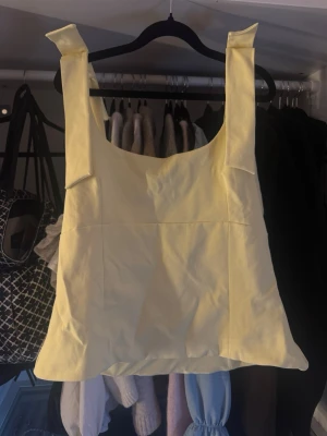 Gul linnetopp med breda axelband - Snygg ljusgul linnetopp med breda axelband och fyrkantig ringning. Toppen har dekorativa knyt på axlarna och en lätt croppad passform. 