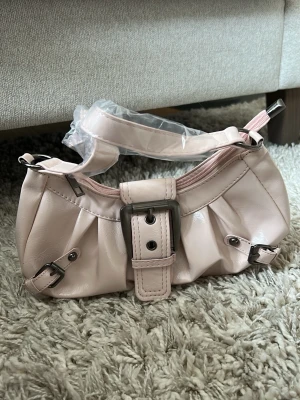 Ljusrosa axelväska med spänne - Söt ljusrosa handväska i glansigt syntetmaterial med silverfärgade metalldetaljer och dekorativa spännen framtill. Väskan har ett kort axelband och dragkedja upptill. Perfekt för att ge din outfit en trendig touch. Ej äkta läder, plast-ish material.