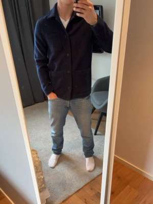 Mörkblå overshirt med fickor - Snygg mörkblå jeansjacka med klassisk krage och stora knappar framtill. Jackan har två stora fickor på framsidan och raka ärmar för en clean look. Perfekt att slänga över en t-shirt för en avslappnad stil. Jag är 183cm och den är lite för liten. Knappt använd