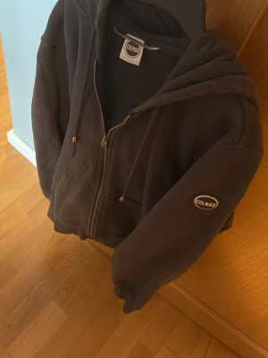 Mörkgrå hoodie från Colmar - Mörkgrå/ lilla mörk  hoodie från Colmar med dragkedja framtill, två fickor och justerbar huva med snören.. Colmar-logga på ärmen  storlek s/m 