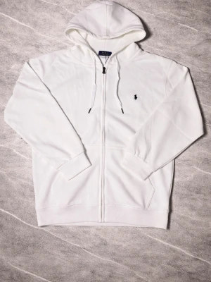 Vit hoodie från Polo Ralph Lauren - Snygg vit hoodie från Polo Ralph Lauren med dragkedja framtill och klassisk huva med dragsko. Aldrig använd och i perfekt skick! Liten broderad logga på bröstet och två fickor fram. Tillverkad i mjukt material som känns skönt mot huden. Perfekt för en clean och stilren look.