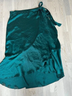 Grön omlottkjol i satin - Snygg grön omlottkjol i glansigt satinliknande material. Kjolen har knytband i midjan och en lätt vågig nederkant som ger en elegant look. Perfekt för dig som vill ha en trendig och bekväm kjol med fin passform. Rök och djurfritt hem. Pruta gärna 🌸