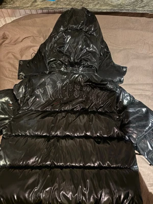 Svart glansig pufferjacka Trapstar - Säljer en svart glansig pufferjacka från Trapstar med stor huva och dragkedja framtill. Jackan har Trapstar-loggan broderad på ryggen och en rund patch på bröstet. Tillverkad i ett blankt, vattenavvisande material som håller dig varm under kalla dagar. Den har en liten repa på huvan.