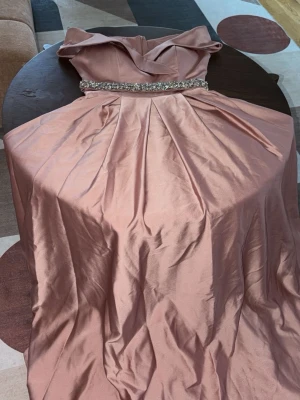 Lång rosa balklänning med pärldetalj - Elegant långklänning i ljusrosa med axelbandslös design och vacker pärlbesatt midja. Klänningen har en utsvängd kjol med veck och är tillverkad i ett glansigt material som ger en lyxig känsla. Perfekt för bal eller festliga tillfällen.