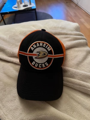 Anaheim Ducks keps från Adidas - Snygg Anaheim Ducks keps från Adidas i svart och orange med broderad logga framtill. Klassisk böjd skärm och justerbar baksida. Perfekt för dig som gillar hockey och vill ha en sportig look. Materialet är bomullsmix.