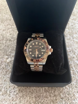 Seiko GMT Klocka “Rootbeer” 40mm – NH34 Automatisk Mod - Säljer en helt nybyggd GMT-klocka i klassisk “Rootbeer” - design. En stilren och unik klocka med premium känsla och noggrant utvalda komponenter.  Specifikationer: 	•	Storlek: 40mm 	•	Urverk: NH34 (automatiskt med GMT-funktion) 	•	Material: 904L rostfritt stål (two-tone rosé/silver) 	•	Glas: Safirglas med datumförstoring 	•	Bezel: Rootbeer-färger 	•	Tavla: Svart med rosé detaljer och datum 	•	Armband: Jubilee (two-tone) med viklås, justerbart 