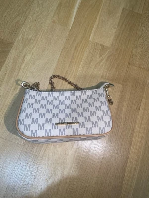 Beige kuvertväska från Michael Kors - Snygg beige kuvertväska från Michael Kors med M-mönster över hela väskan. Väskan har guldfärgad kedja som handtag och dragkedja upptill. Tillverkad i tåligt material med ljusbruna detaljer längs kanterna.