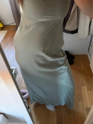 Ljusgrön långklänning i satin - Säljer en elegant ljusgrön långklänning i satin med smala axelband och öppen rygg. Klänningen har en mjuk, glansig yta och följsam passform som ger en snygg siluett. Perfekt för dig som vill ha en stilren och trendig look.