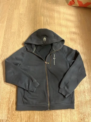 Svart hoodie från Sail Racing - Mörkblå hoodie från Sail Racing med dragkedja framtill och en extra bröstficka med dragkedja. Hoodien har broderad logga på bröstet och detaljer vid huvan. Tillverkad i mjukt och värmande material, perfekt för chill dagar.