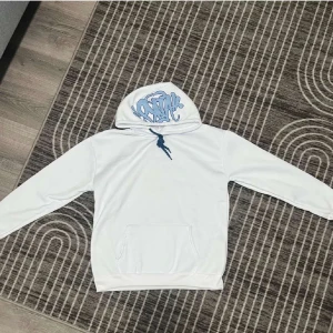 Vit hoodie med blått tryck - Säljer en vit hoodie med blått graffiti-inspirerat tryck på huvan och blå snörning. Tröjan har en klassisk känguruficka framtill och långa ärmar. Perfekt för dig som gillar streetwear och vill ha en clean look med en twist.