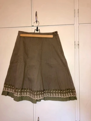 Olivgrön kjol med spetsdetaljer - Säljer en olivgrön kjol i A-linjeform med bred volangkant och dekorativ spets i beige längs nederkanten. Kjolen har en mjuk struktur och är tillverkad i ett lätt tyg, perfekt för dig som gillar boho-stil.