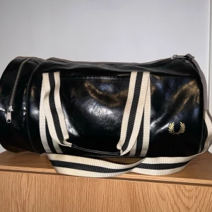 Fred Perry Barrel Bag - Klassisk duffelväska från Fred Perry i svart med vita randiga remmar och guldfärgad logga. • Perfekt som gymväska eller weekendbag • Sparsamt använd, endast ett märke som syns på bilden