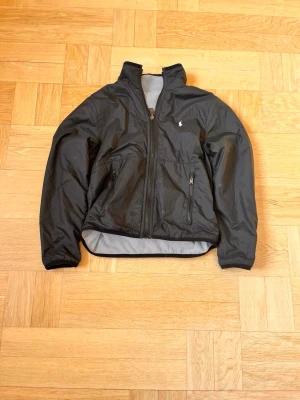 Svart vindjacka från Polo Ralph Lauren - Snygg svart vindjacka från Polo Ralph Lauren med dragkedja framtill och två fickor med dragkedja. Jackan har hög krage och en liten vit logga på bröstet. Tillverkad i lätt och tåligt material med meshfoder på insidan.