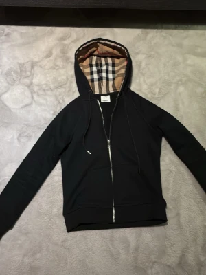 Svart hoodie från Burberry med dragkedja - Snygg svart hoodie från Burberry med dragkedja framtill och klassiskt rutigt foder i huvan. Hoodien har dragsko i huvan och diskret Burberry-logga broderad på bröstet. Perfekt för dig som gillar stilrena plagg med exklusiva detaljer.