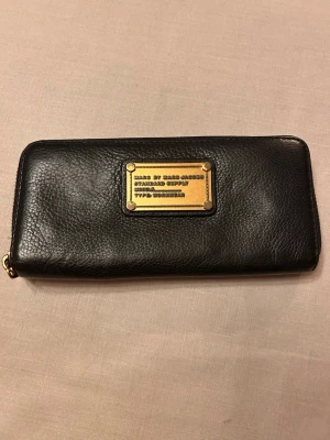 Svart plånbok från Marc by Marc Jacobs - Snygg svart plånbok i äkta läder från Marc by Marc Jacobs. Plånboken har guldfärgade metalldetaljer, dragkedja runtom och flera fack för kort och sedlar. 20x10cm