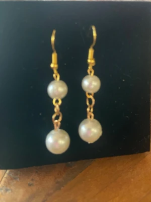 Guldtonade örhängen med pärlor - Snygga hängande örhängen med två vita pärlor på varje örhänge och guldfärgade krokar och kedjor. Klassisk design som ger en elegant touch till din stil. Perfekta för dig som gillar tidlösa accessoarer med pärldetaljer.