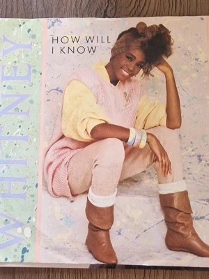 Whitney Houston vinylskiva - Säljer en retro vinylskiva med Whitney Houston. Omslaget har ett färgstarkt, stänkmålat mönster i grönt, blått och vitt samt ett foto på artisten i vit klänning. Skivan innehåller låtar som 'How Will I Know' och 'Someone For Me'. Perfekt för samlare av klassisk popmusik.