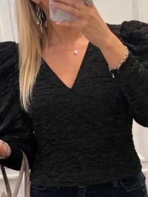 Svart topp med puffärmar och v-ringning - Trendig svart topp med markerade puffärmar och snygg v-ringning. Materialet har en strukturerad, lite skrynklig look som ger extra edge. Passformen är normal och toppen är croppad, perfekt till höga jeans eller kjol.