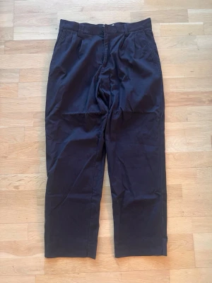 Svarta chinos med raka ben - Snygga svarta chinos med raka ben och en pösig passform upptill. Byxorna har bälteshällor, två sidofickor fram och två bakfickor med knapp. Byxorna är gjorda av bomull och är helt oanvända. 