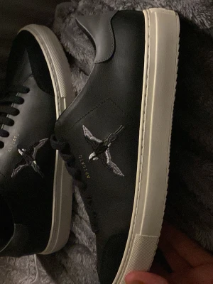 Svarta Axel Arigato sneakers med broderad fågel - Snygga svarta sneakers från Axel Arigato med vit sula och broderad fågel på sidan. Skorna har klassisk låg profil, svarta skosnören och är tillverkade i skinn. Perfekta för dig som gillar stilrena detaljer och streetwear vibes.