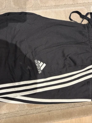 Svarta Adidas mjukisbyxor L - Klassiska svarta mjukisbyxor från Adidas med vita ränder längs benen och logga på sidan. Byxorna har dragsko i midjan och är tillverkade i 70% bomull och 30% polyester. Perfekta för chill eller träning.