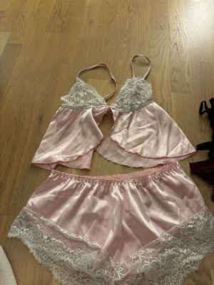 Rosa satin pyjamasset med spetsdetaljer - Sött pyjamasset i ljusrosa satin med vita spetsdetaljer. Toppen har tunna axelband, spets över bysten och en liten rosett framtill. Shortsen har elastisk midja och spetskant nedtill. Helt ny, tvättad. 