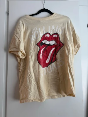 Rolling Stones beige t-shirt - Säljer en beige t-shirt med det ikoniska Rolling Stones-trycket framtill, stor röd tunga och vita bokstäver. T-shirten har rund hals och korta ärmar. Perfekt för dig som gillar bandmerch och vill ha en avslappnad stil.