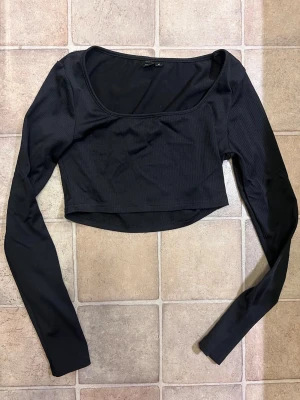 Svart croppad långärmad topp - Trendig svart croppad topp med långa ärmar och rundad urringning. Tillverkad i ett mjukt, ribbat material som ger en snygg och bekväm passform. Perfekt att styla med höga jeans eller kjol för en cool look.