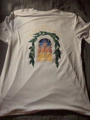 Vit t-shirt med tryck - Perfekt för sommaren. Vit t-shirt från Casa/.      blanca med färgstarkt tryck av en byggnad och gröna blad både fram och bak. T-shirten har rund hals och korta ärmar. Perfekt för dig som gillar stilrena plagg med unika detaljer.