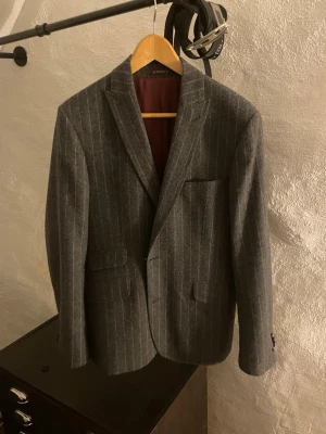 Grå Morris kavaj med kritstrecksrand - Snygg grå kavaj med kritstrecksrand och klassisk passform. Kavajen har två framfickor med lock, bröstficka och två knappar vid ärmslutet. Fodret är vinrött och materialet ser ut att vara ullblandning. Perfekt för en stilren look. Nypris ca 5000kr