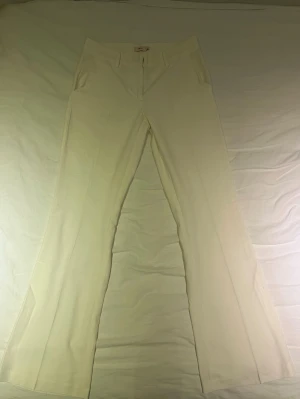 Vida vita kostymbyxor - Snygga vita kostymbyxor med utsvängda ben och hög midja. Byxorna har fickor fram och klassisk dragkedja. Perfekta för en clean och stilren look. Materialet ser ut att vara ett mjukt tyg med lite stretch.