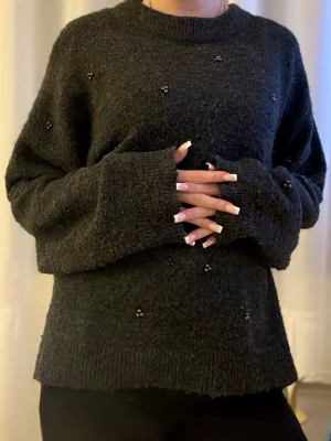 Mörkgrå stickad tröja med pärlor - Mysig mörkgrå stickad tröja med små pärldetaljer utspridda över hela tröjan. Tröjan har en avslappnad passform, rund halsringning och ribbade muddar vid ärmslut och nederkant. Perfekt för kalla dagar och enkel att styla till olika outfits.