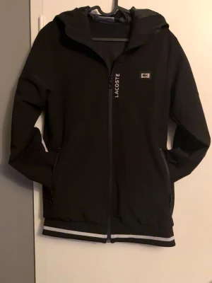 Svart vindjacka från Lacoste med dragkedja - Svart vindjacka från Lacoste med hel dragkedja, huva och vita detaljer på ärmar och huva. Märket syns tydligt på bröstet och längs dragkedjan. Två fickor med dragkedja framtill och ribbade muddar nedtill. Perfekt för en sportig och stilren look.