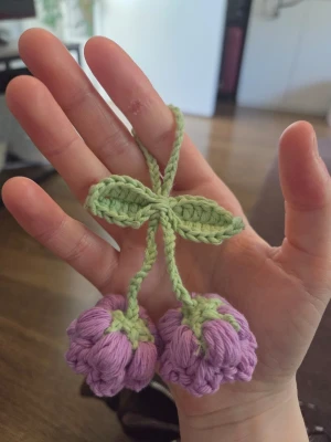 Virkad nyckelring med lila blommor - Handgjord virkad accessoar i form av två lila blommor med gröna blad och stjälkar. Perfekt som dekoration till väska eller nyckelknippa. Tillverkad i mjukt garn och har en söt, lekfull design.