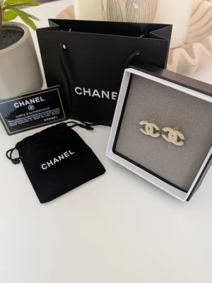 ChaneI örhängen - Exklusiva örhängen i form av den ikoniska, sammanflätade logotypen, presenterade med kompletta tillbehör. ​Design: Örhängena har en tidlös design i en ljust guldfärgad eller krämvit finish med en slät, elegant yta. ​Tillbehör: Säljes som ett set med tillhörande svart originalask, en liten skyddande sammetspåse och en svart bärkasse i papper. ​