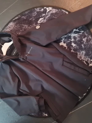 Svart softshelljacka från Sail Racing - Snygg svart softshelljacka från Sail Racing med minimalistisk design. Jackan har diskreta knappar och är tillverkad i ett lätt, vindtätt material som passar perfekt för blåsiga dagar. Klassisk passform och stilren look. En liten lagning vid fickan som inte stör eller påverkar funktionen. Stl 170.