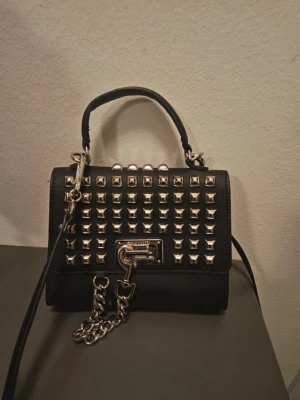 Steve Madden Svart handväska med nitar och kedja - Steve Madden. Cool svart handväska i rektangulär form med silverfärgade nitar på framsidan och en dekorativ kedja. Väskan har både handtag och lång axelrem, samt ett metallspänne framtill. Perfekt för dig som gillar en edgy och trendig stil.