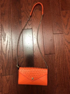 Rebecca Minkoff Orange axelremsväska med nitar - Rebecca Minkoff. Snygg orange kuvertväska i läder med guldfärgade nitar och kedjerem. Väskan har ett kuvertformat lock med tryckknapp och coola detaljer som ger en edgy vibe. Perfekt för dig som vill sticka ut med accessoarerna.