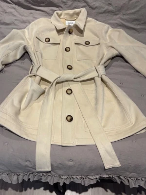 Beige kappa med bälte från isolde - Beige kappa från isolde med stora bruna knappar, två bröstfickor och ett matchande knytbälte i midjan. Inga tecken på användning 💕