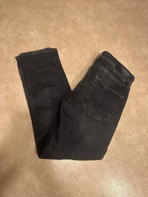 Gråa jeans från Jack & Jones  - Snygga svarta jeans från Jack & Jones 30/30 med klassisk femficksdesign och svarta sömmar. Jeansen har en diskret tvättad look och är tillverkade i stretchigt denim för extra komfort. Perfekta för en stilren och avslappnad outfit.