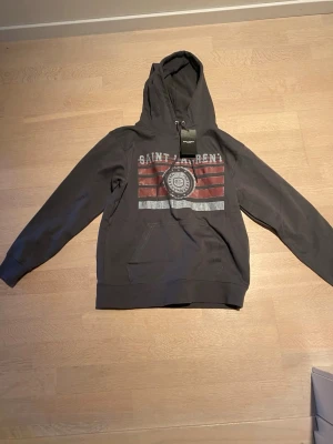 Grå Saint Laurent hoodie med tryck - Snygg grå hoodie från Saint Laurent med huva och dragsko. Framsidan har ett stort tryck med texten 'SAINT LAURENT' och röda samt vita ränder. Klassisk känguruficka framtill och ribbade muddar. Perfekt för en avslappnad streetwear-look. 